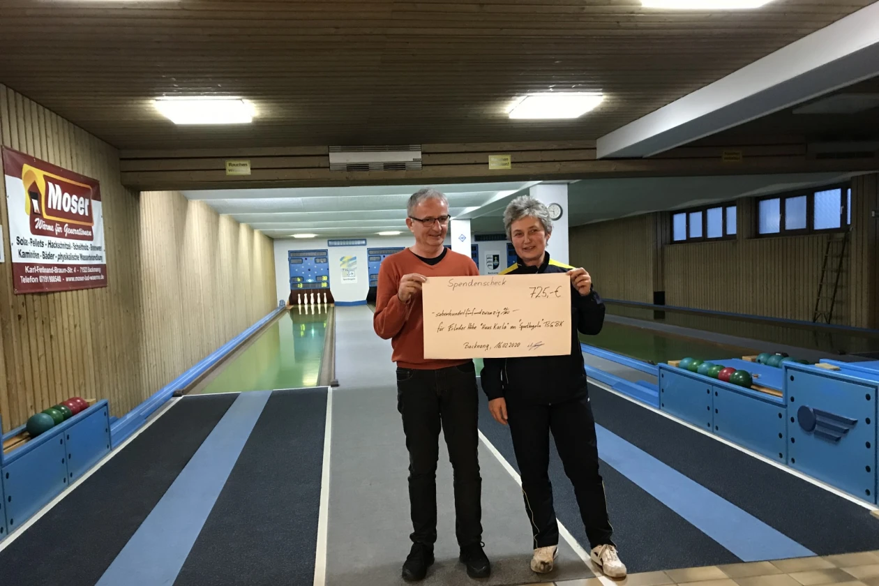 Zwei Personen stehen auf einer Kegelbahn. Beide halten gemeinsam einen Scheck in der Hand. An der Wand h&auml;ngen Bilder und an der Decke h&auml;ngen leuchtende Lampen.