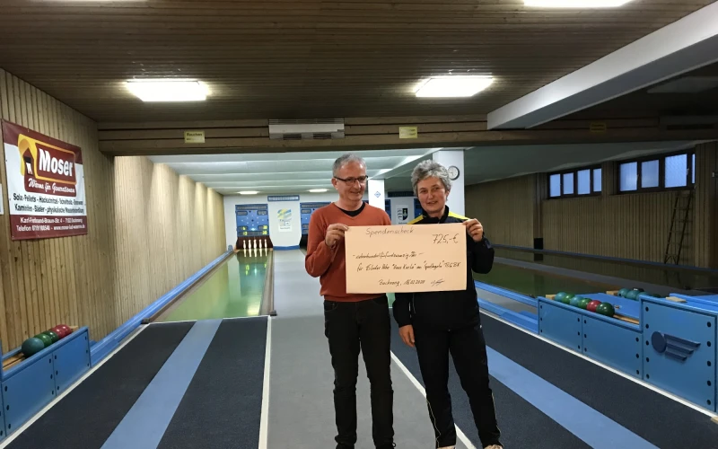 Zwei Personen stehen auf einer Kegelbahn. Beide halten gemeinsam einen Scheck in der Hand. An der Wand h&auml;ngen Bilder und an der Decke h&auml;ngen leuchtende Lampen.