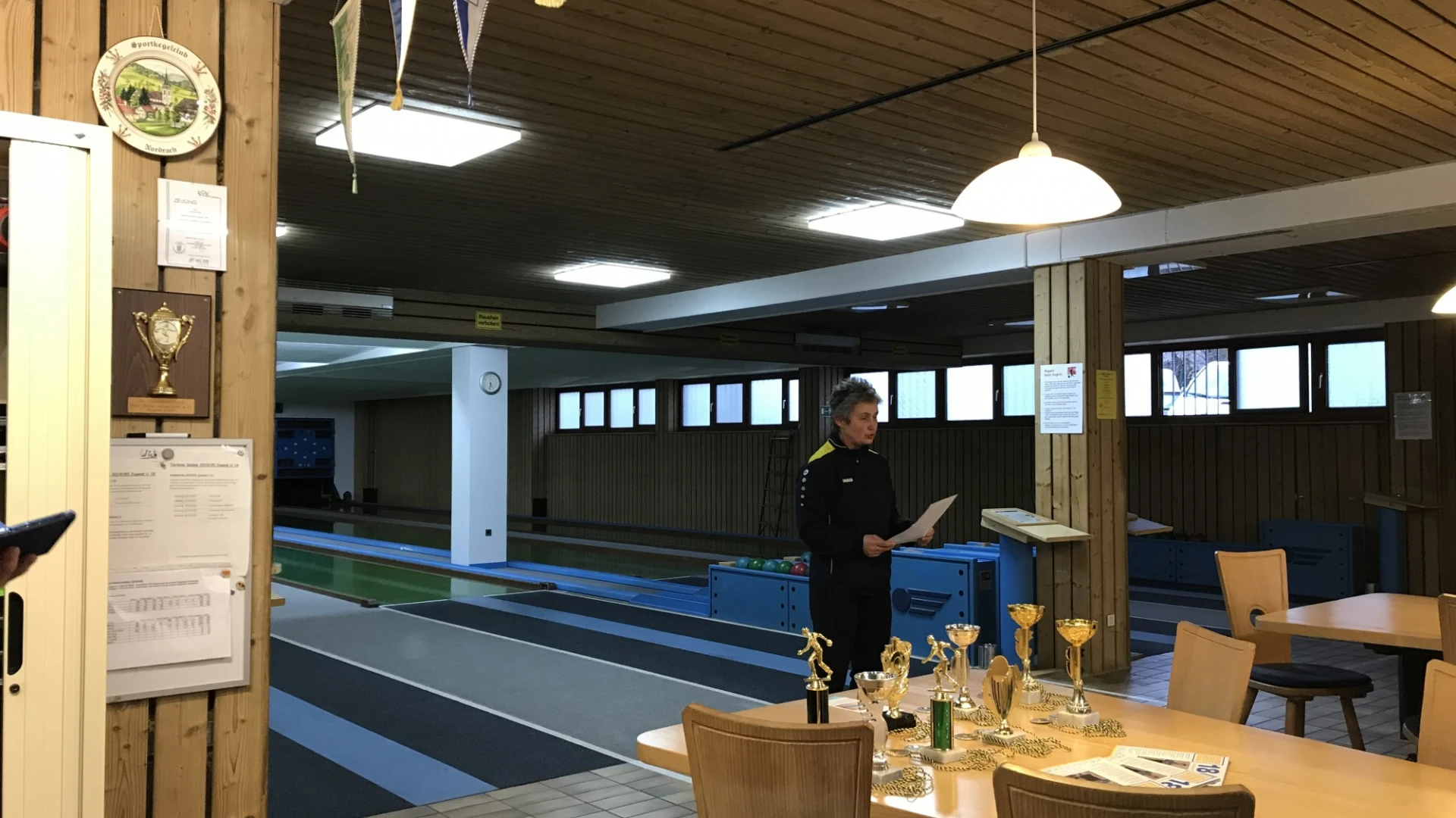 Innenaufnahme einer Kegelbahn. Im Vordergrund steht ein Tisch mit mehreren Pokalen. Eine Frau h&auml;lt ein Blatt Papier in der Hand.