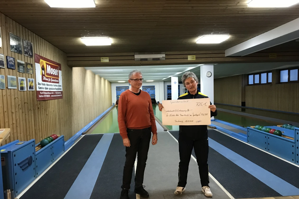Zwei Personen stehen auf einer Kegelbahn. Die Person rechts h&auml;lt einen Scheck in der Hand. An der Wand h&auml;ngen Bilder und an der Decke h&auml;ngen leuchtende Lampen.