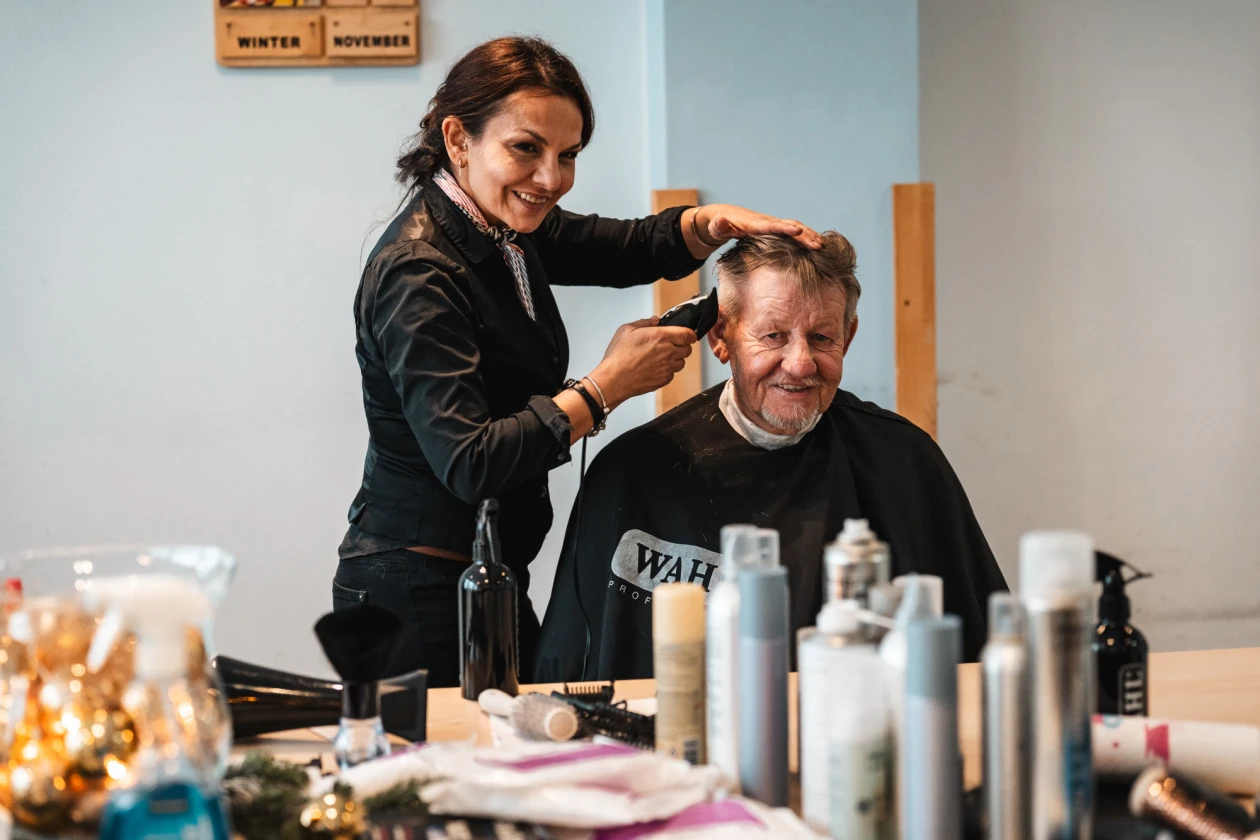 Eine Friseurin schneidet die Haare eines &auml;lteren Mannes in einem hellen Raum. Auf dem Tisch vor ihm sind verschiedene Friseurprodukte und Beh&auml;lter angeordnet.