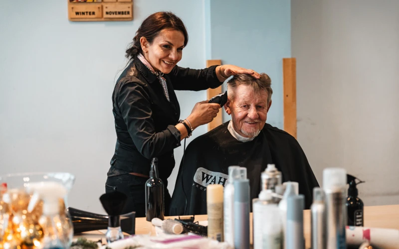 Eine Friseurin schneidet die Haare eines &auml;lteren Mannes in einem hellen Raum. Auf dem Tisch vor ihm sind verschiedene Friseurprodukte und Beh&auml;lter angeordnet.