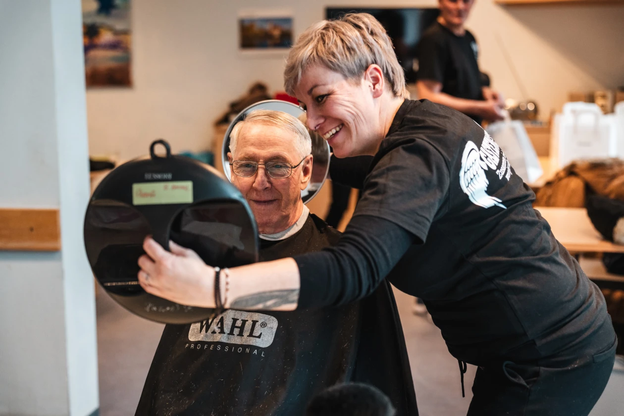 Eine Friseurin schneidet einem &auml;lteren Mann mit Brille die Haare. Im Hintergrund sind weitere Personen in einem hellen Raum zu sehen.