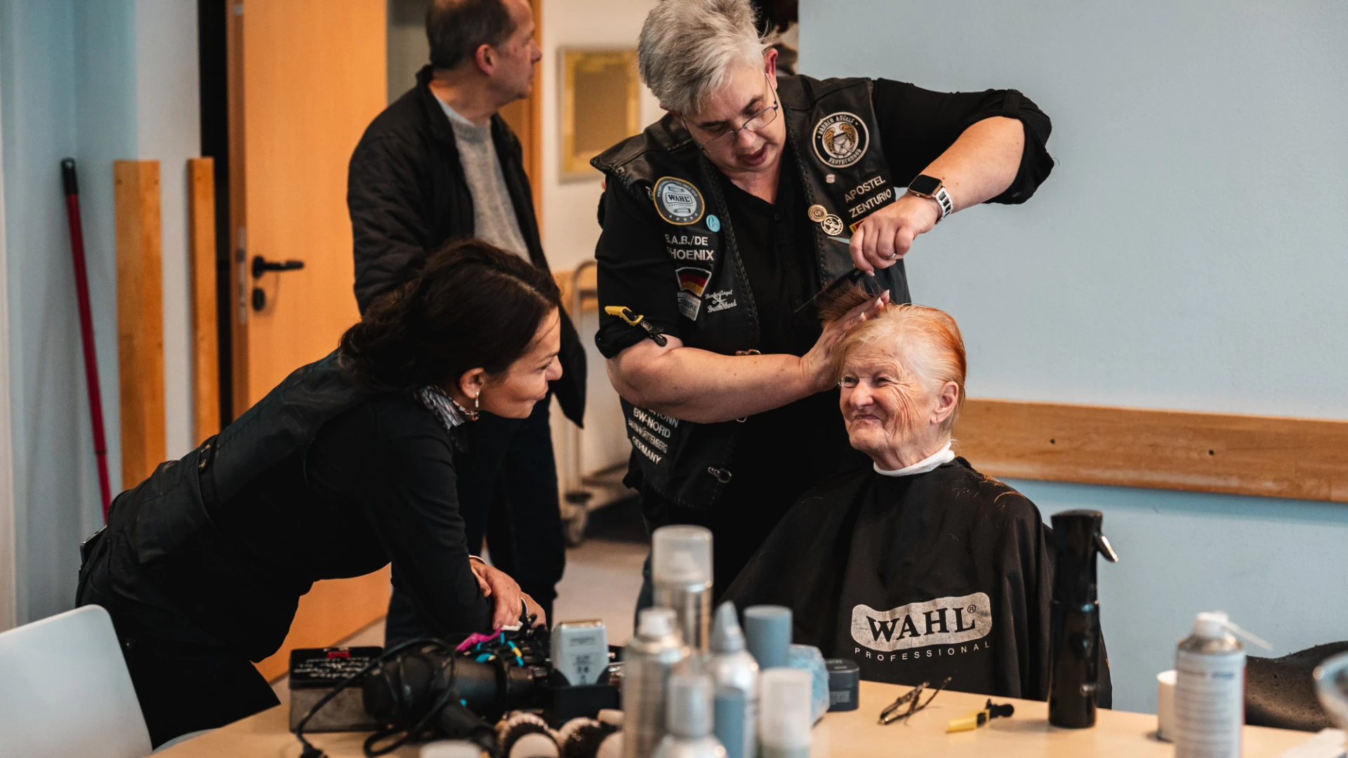 Eine Friseurin in schwarzer Weste mit Abzeichen schneidet einer &auml;lteren Frau die Haare in einem hellen Raum.