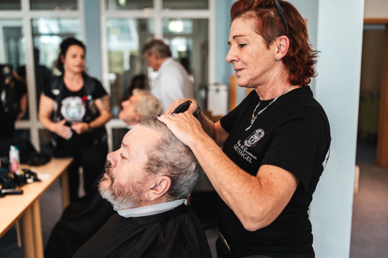 Eine Friseurin mit r&ouml;tlichen Haaren schneidet einem &auml;lteren Mann mit grauem Bart die Haare. Im Hintergrund sind weitere Personen in einem hellen Raum zu sehen.