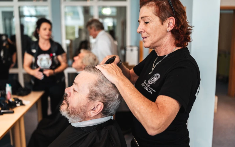 Eine Friseurin mit r&ouml;tlichen Haaren schneidet einem &auml;lteren Mann mit grauem Bart die Haare. Im Hintergrund sind weitere Personen in einem hellen Raum zu sehen.