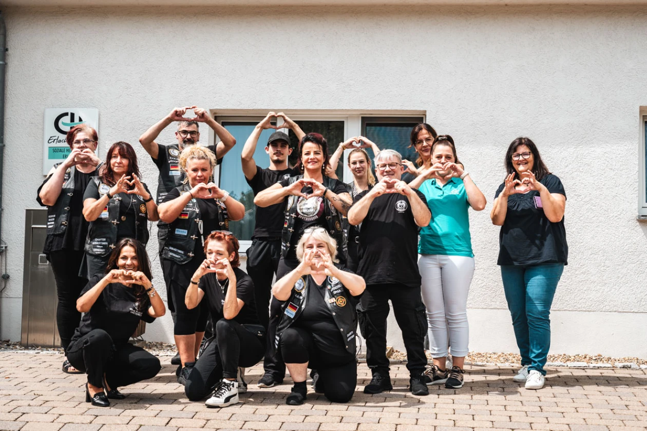 Eine Gruppe von etwa 15 Personen in schwarzen T-Shirts und einer Person in T&uuml;rkis steht vor einer wei&szlig;en Wand und formt mit ihren H&auml;nden &uuml;ber ihren K&ouml;pfen ein Herzzeichen