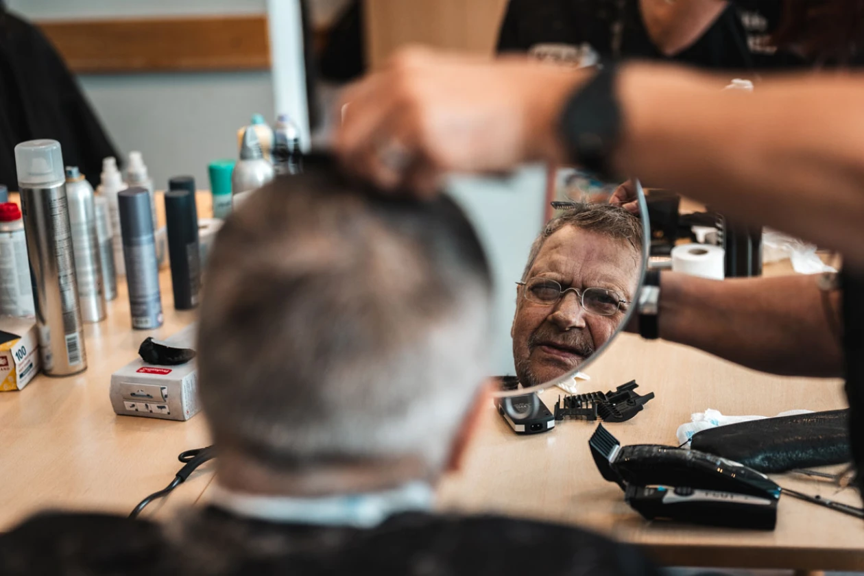 Eine Friseurin schneitet einem Mann mit schwarzem Haar die Haare. Verschiedene Haarpflegeprodukte stehen auf dem Tisch, w&auml;hrend weitere Personen im Hintergrund arbeiten.