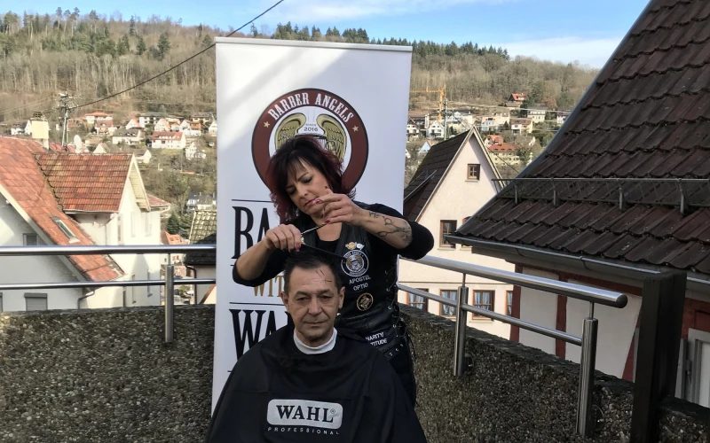 Ein Friseurin schneidet einem Mann auf einem Balkon mit Blick auf H&auml;usern und Wald im Hintergrund die Haare. Er tr&auml;gt einen Frisierumhang.