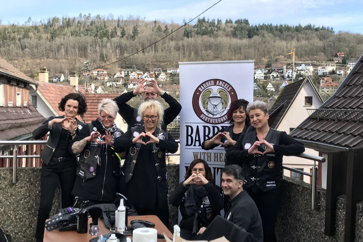 Acht Personen in schwarzer Kleidung posieren zusammen auf einem Dach mit einer Barber-Werbefahne. Im Hintergrund ist ein Wohngebiet und Wald zu sehen. Die Gruppe macht herzf&ouml;rmige Gesten mit den H&auml;nden.