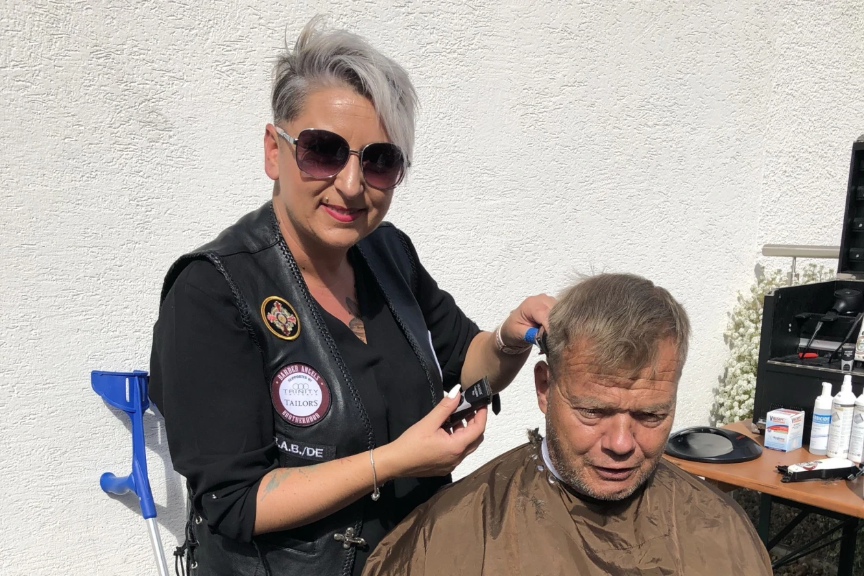 Eine Frau in schwarzer Kleidung schneidet einem Mann die Haare. In ihrer rechten Hand h&auml;lt sie eine Haarschneidemaschine. Der Mann tr&auml;gt einen braunen Umhang mit der Aufschrift &sbquo;TAILORS&lsquo;. Die Frau tr&auml;gt eine Sonnenbrille und steht w&auml;hrend des Haarschnitts hinter dem Mann. Im Hintergrund sind eine wei&szlig;e Wand, eine Kr&uuml;cke sowie weitere Materialien auf einem Tisch zu sehen.