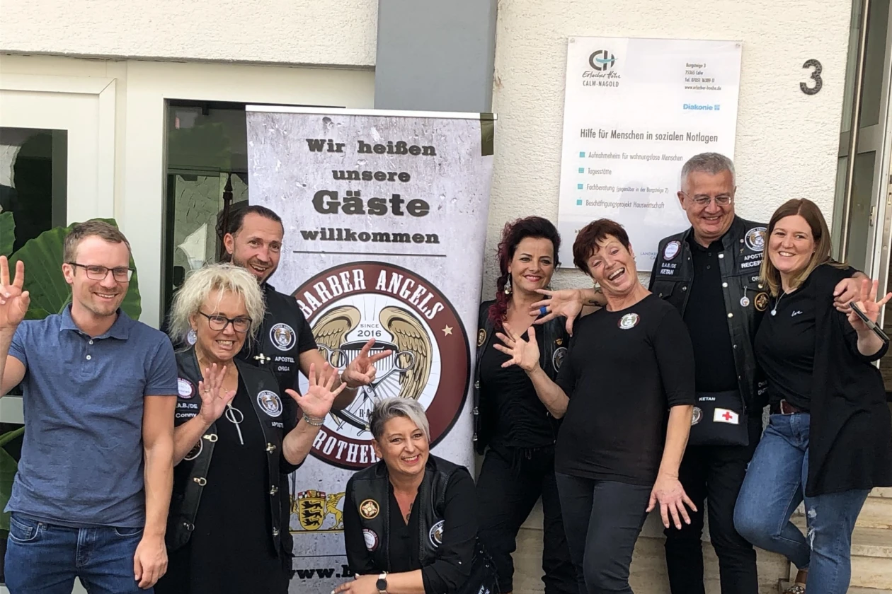 Gruppe von acht Personen, die vor einem Banner mit der Aufschrift "Wir hei&szlig;en unsere G&auml;ste willkommen" stehen. Sie stehen vor einem Geb&auml;ude.