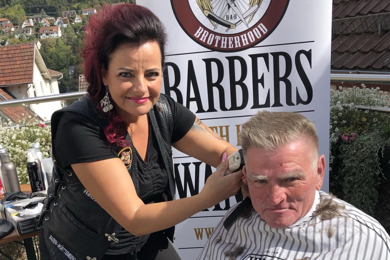 Eine junge Frau in schwarzer Kleidung schneidet im Freien einem Mann mit grauem Haar die Haare. Im Hintergrund sind H&auml;user, B&auml;ume und ein Banner zu sehen, auf dem die Worte "BROTHERHOOD" und &sbquo;BARBERS&lsquo; erkennbar sind.