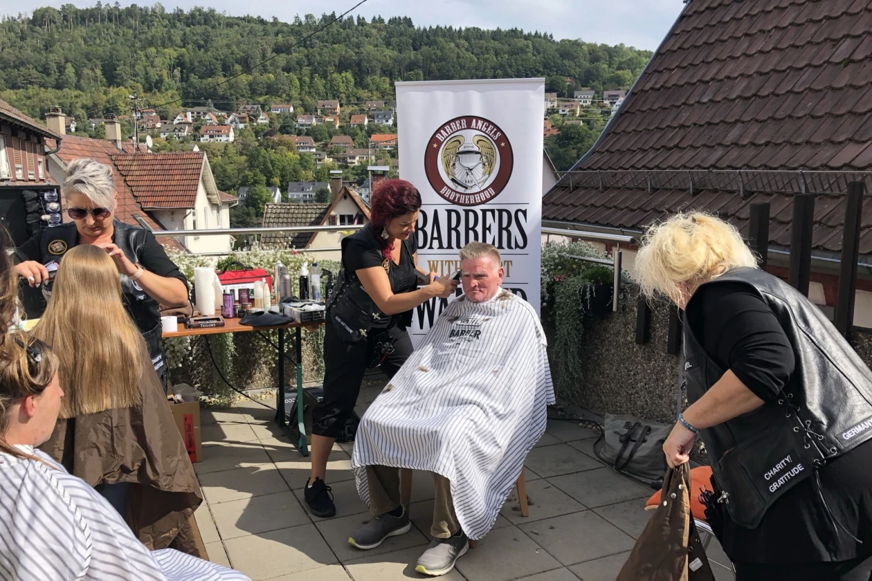 Auf der Terrasse sind zwei Frauen zu sehen, die zwei Personen die Haare schneiden. Eine weitere Person sitzt wartend mit Umgang da und eine schwarz gekleidete Frau holt etwas vom Boden. Im Hintergrund befindet sich ein Banner sowie eine Stadtsiedlung umrahmt von Wald.