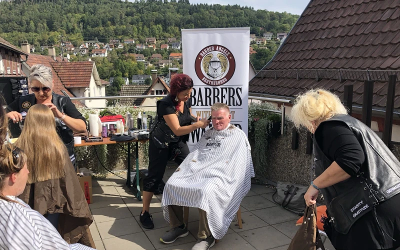 Auf der Terrasse sind zwei Frauen zu sehen, die zwei Personen die Haare schneiden. Eine weitere Person sitzt wartend mit Umgang da und eine schwarz gekleidete Frau holt etwas vom Boden. Im Hintergrund befindet sich ein Banner sowie eine Stadtsiedlung umrahmt von Wald.