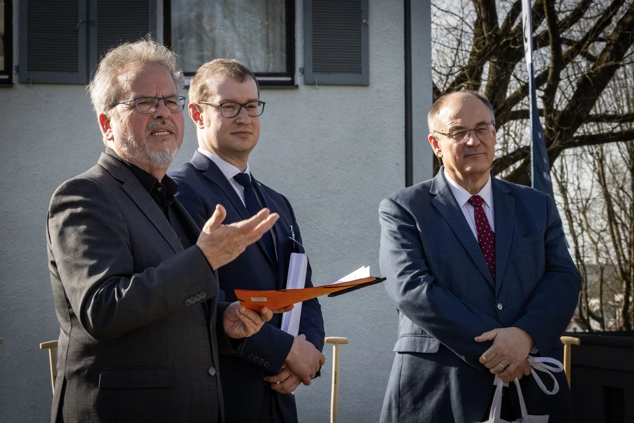 Drei M&auml;nner stehen vor einem Geb&auml;ude. Der Mann links spricht und gestikuliert mit einer Hand, w&auml;hrend er in der anderen Hand eine Mappe h&auml;lt. Der Mann in der Mitte tr&auml;gt Unterlagen in den H&auml;nden und der Mann rechts h&auml;lt eine Tragetasche.