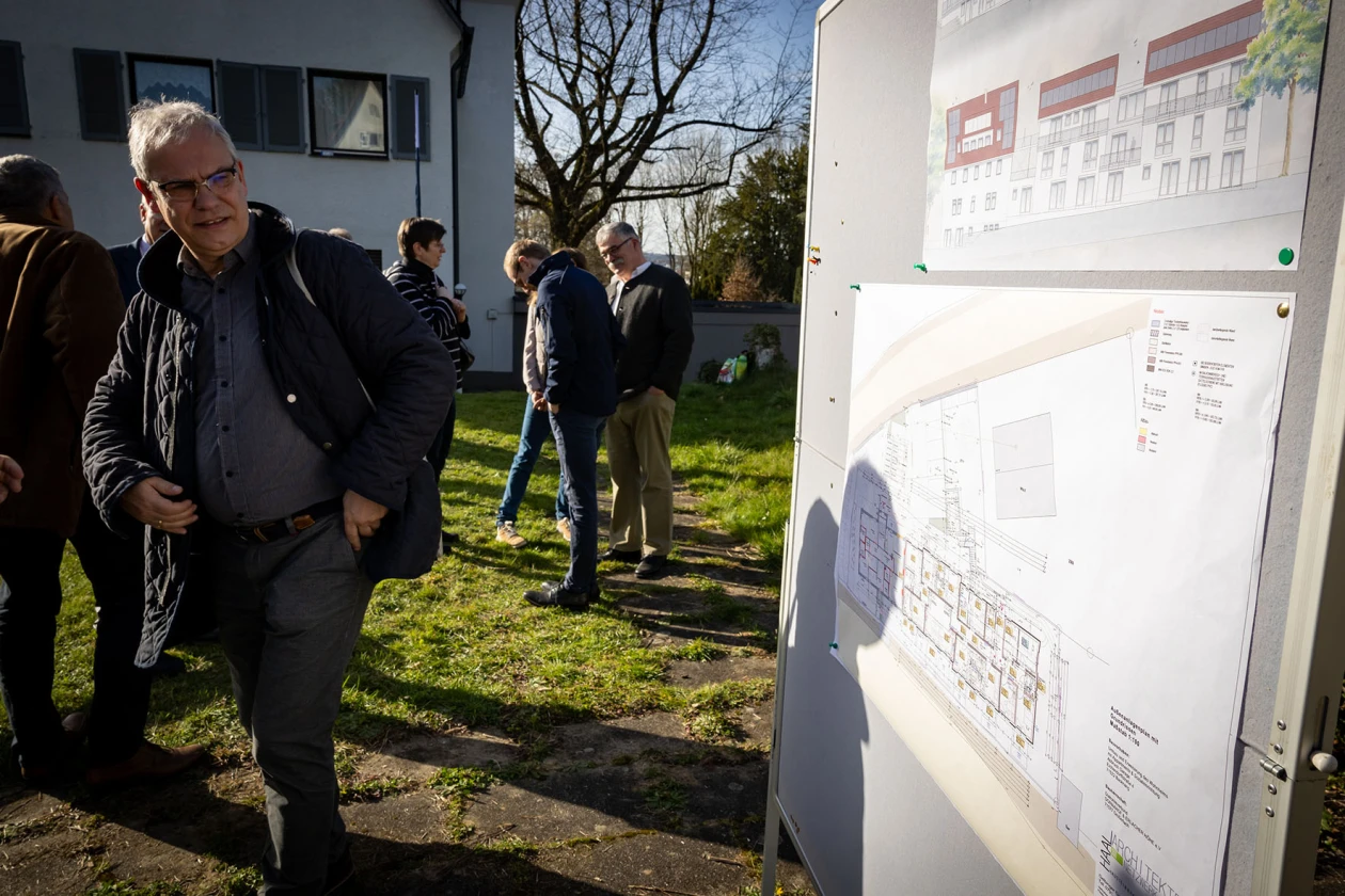 Mehrere Personen stehen auf einer Wiese und unterhalten sich. Rechts im Bild ist eine Informationstafel mit einem Bauplan zu sehen. Im Hintergrund ist ein Geb&auml;ude mit kahlen B&auml;umen zu sehen.