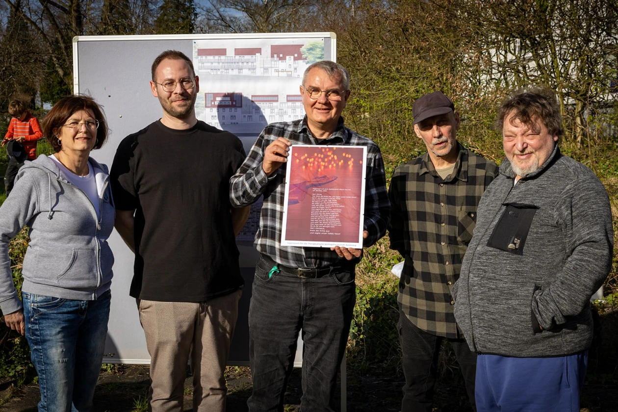 F&uuml;nf Personen stehen im Freien vor einer Informationstafel mit Bauplan und blicken in die Kamera. Der Mann in der Mitte h&auml;lt ein gerahmtes, rotes Dokument in den H&auml;nden.