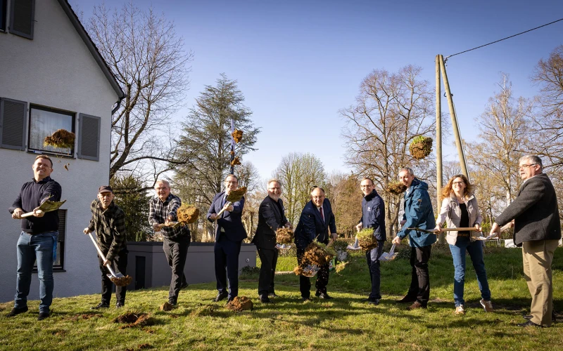 Zehn Personen stehen nebeneinander auf einer Wiese, alle haben einen Spaten in der Hand und werfen l&auml;chelnd Erde in die Luft. Im Hintergrund sind ein Geb&auml;ude, B&auml;ume und ein klarer Himmel zu sehen.