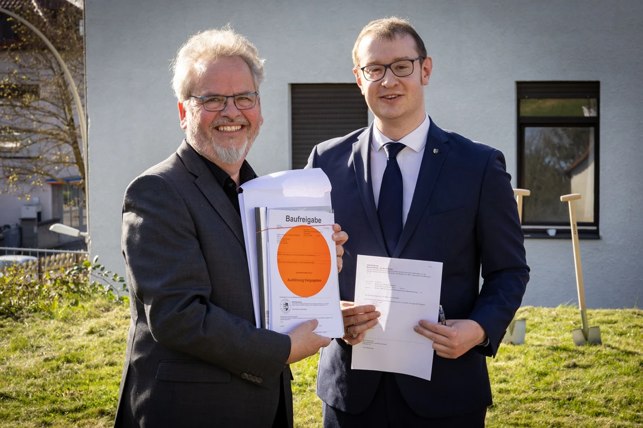 Zwei M&auml;nner stehen mit Anz&uuml;gen auf einer Wiese im Freien vor einem Geb&auml;ude. Der Mann links h&auml;lt freudig eine Urkunde mit einem roten Punkt in die Kamera. Der Mann rechts h&auml;lt ein weiteres Dokument in den H&auml;nden. Im Hintergrund stehen zwei Spaten im Rasen.