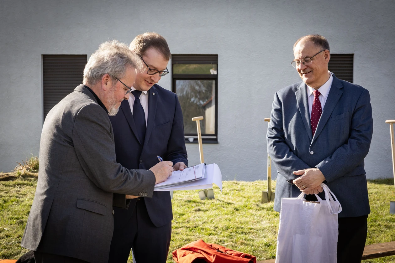 Drei M&auml;nner stehen auf einer Wiese vor einem Geb&auml;ude, alle drei in dunklen Anz&uuml;gen gekleidet. Ein Mann unterschreibt ein Dokument in den H&auml;nden eines anderen Mannes, w&auml;hrend der dritte Mann l&auml;chelnd beobachtet und eine Tasche in den H&auml;nden h&auml;lt.