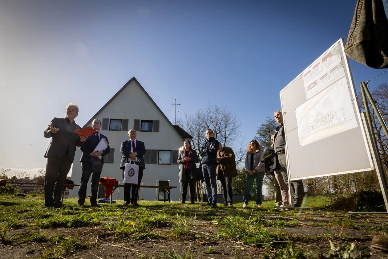 Eine Gruppe von Menschen steht auf einem Grundst&uuml;ck auf einer Wiese vor einem Geb&auml;ude und einer Informationstafel.