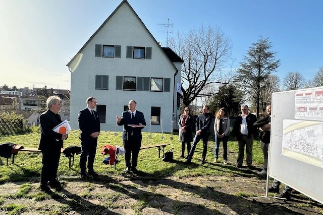 Mehrere Personen stehen im Freien auf einer Wiese vor einem wei&szlig;en, mehrst&ouml;ckigen Haus. Rechts von ihnen befindet sich eine Informationstafel mit Baupl&auml;nen.