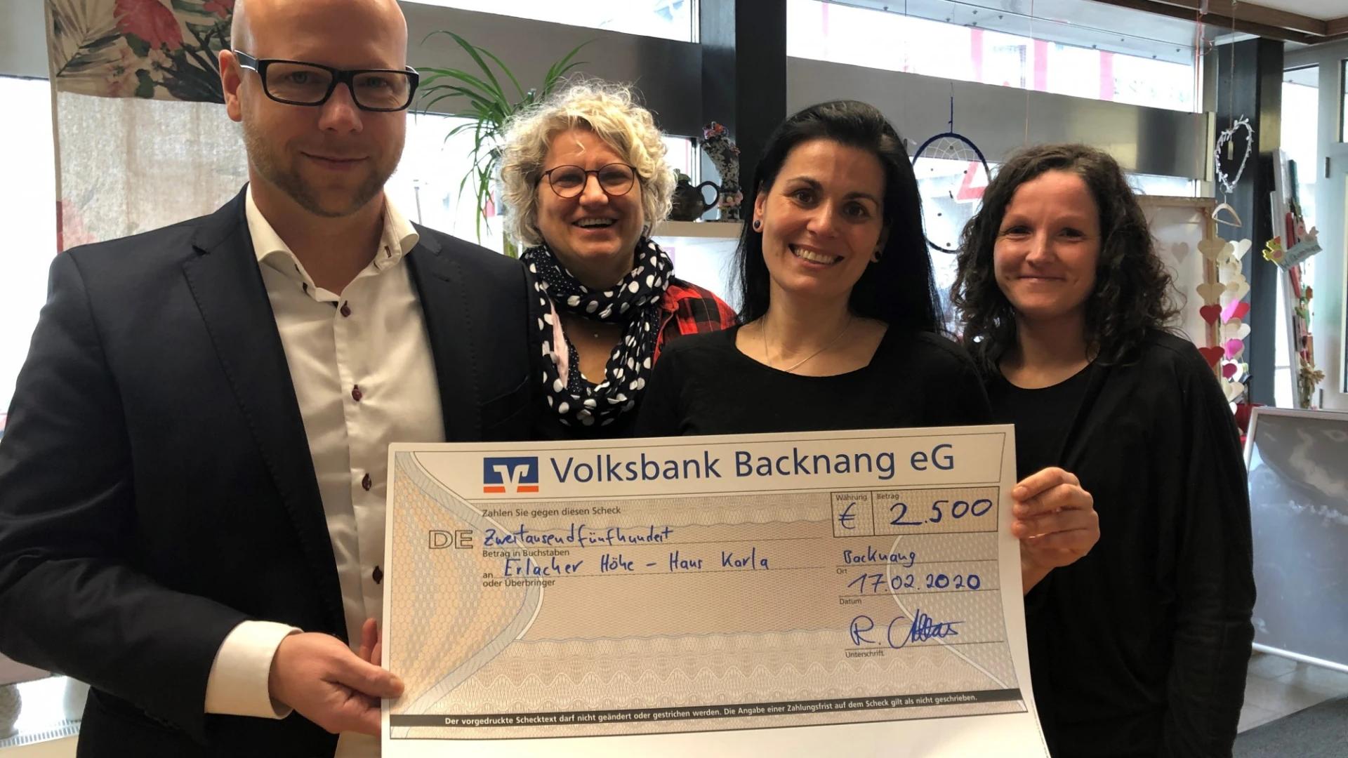 Vier Personen stehen in einem Raum mit Fenster und Dekorationen. Sie halten einen Scheck in der Hand und l&auml;cheln in die Kamera.