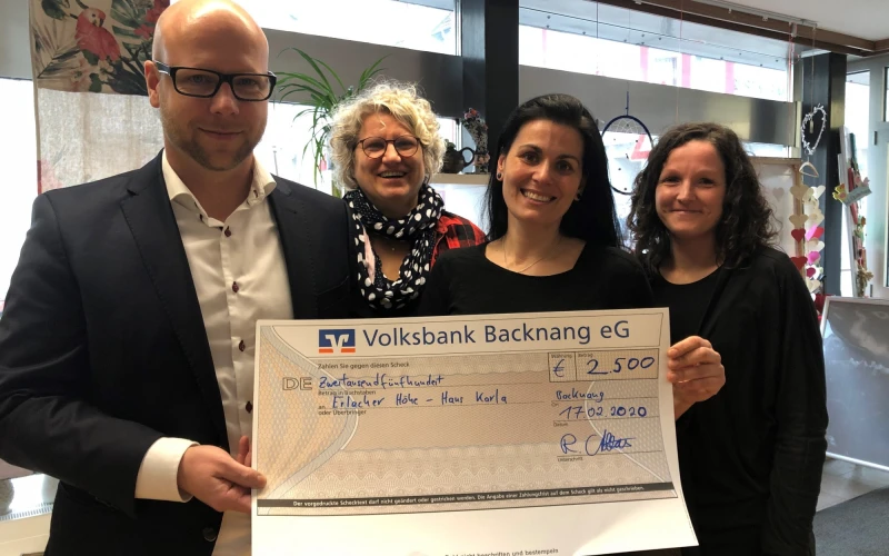 Vier Personen stehen in einem Raum mit Fenster und Dekorationen. Sie halten einen Scheck in der Hand und l&auml;cheln in die Kamera.