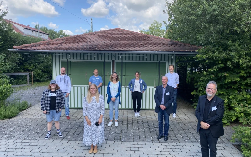 Neun Personen stehen auf einem gepflasterten Boden vor einem kleinen Haus mit gr&uuml;ner Fassade und rotem Ziegeldach, alle l&auml;cheln in die Kamera.