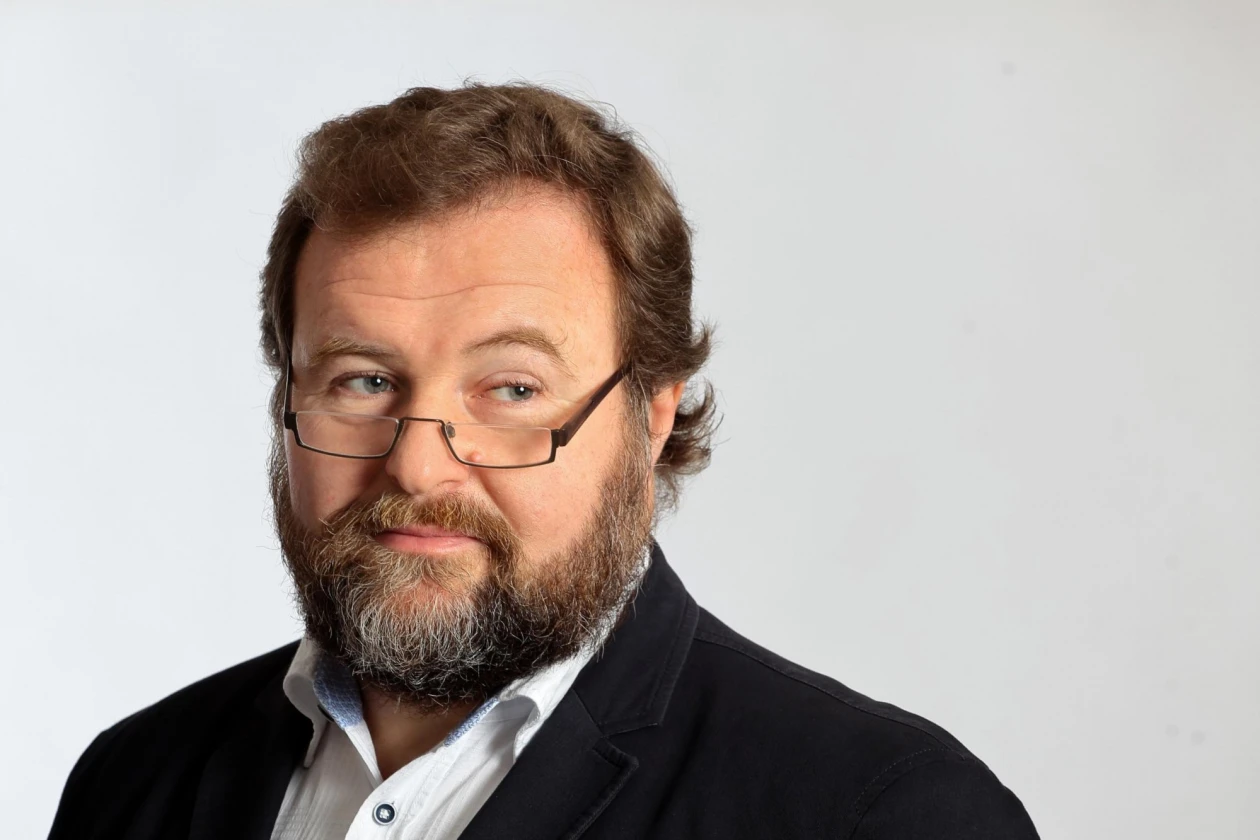 &Auml;lterer b&auml;rtiger Mann mit Brille und schwarzem Sakko und wei&szlig;em Hemd. Er steht vor einem neutralen Hintergrund.