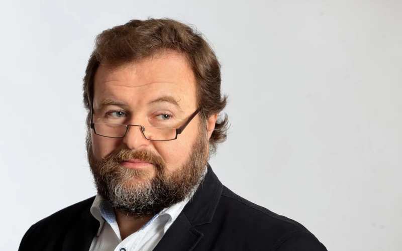 &Auml;lterer b&auml;rtiger Mann mit Brille und schwarzem Sakko und wei&szlig;em Hemd. Er steht vor einem neutralen Hintergrund.