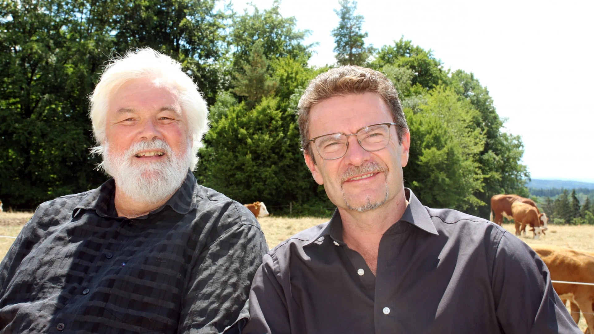 Zwei &auml;ltere M&auml;nner l&auml;cheln in die Kamera, einer hat wei&szlig;es Haar und Bart, der andere dunkle Haare und tr&auml;gt Brille. Im Hintergrund B&auml;ume und eine Weide mit K&uuml;hen. Sie tragen beide dunkle Oberteile und scheinen an einem sonnigen Tag drau&szlig;en zu sein.