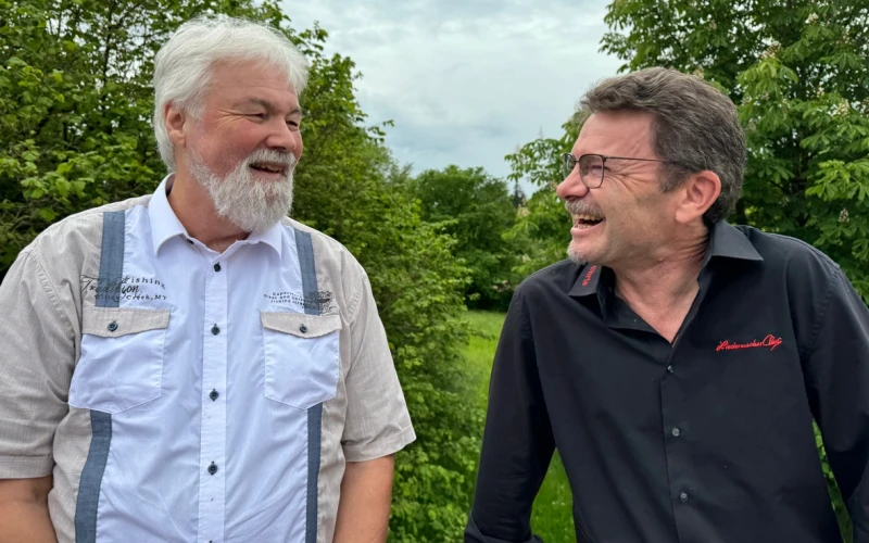 Zwei M&auml;nner stehen sich nebeneinander und l&auml;cheln sich an. Der wei&szlig;haarige Mann tr&auml;gt Bart und hat ein beige-blaues Hemd an. der dunkelhaarige Mann tr&auml;gt einen kurzen Bart, eine Brille und ein schwarzes Hemd mit rotem Stickmuster.