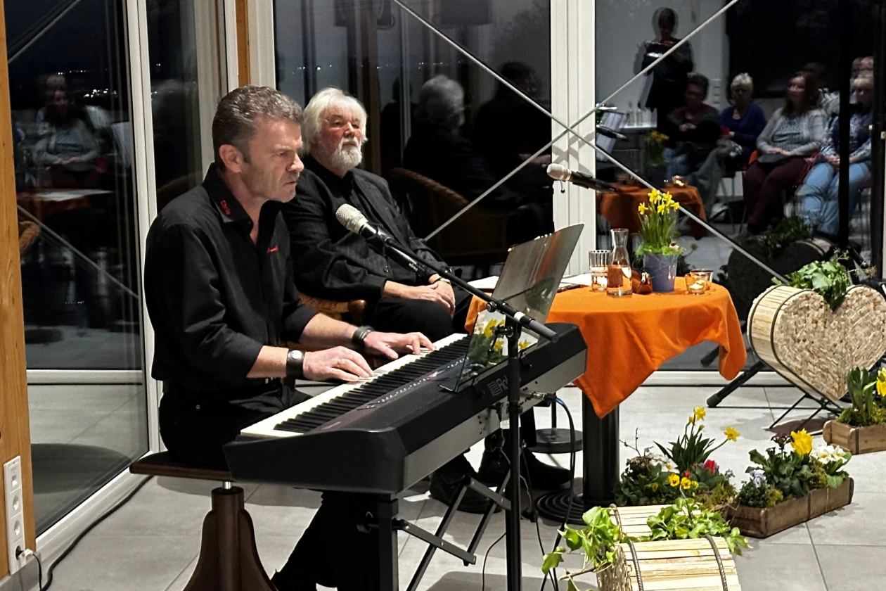 Ein Mann sitzt an einem E-Piano, hinter ihm sitzt ein Mann mit wei&szlig;en Haaren und grauem Bart an einem kleinen runden Tisch. Der Raum hat gro&szlig;e Fenster und ist fr&uuml;hlingshaft dekoriert.