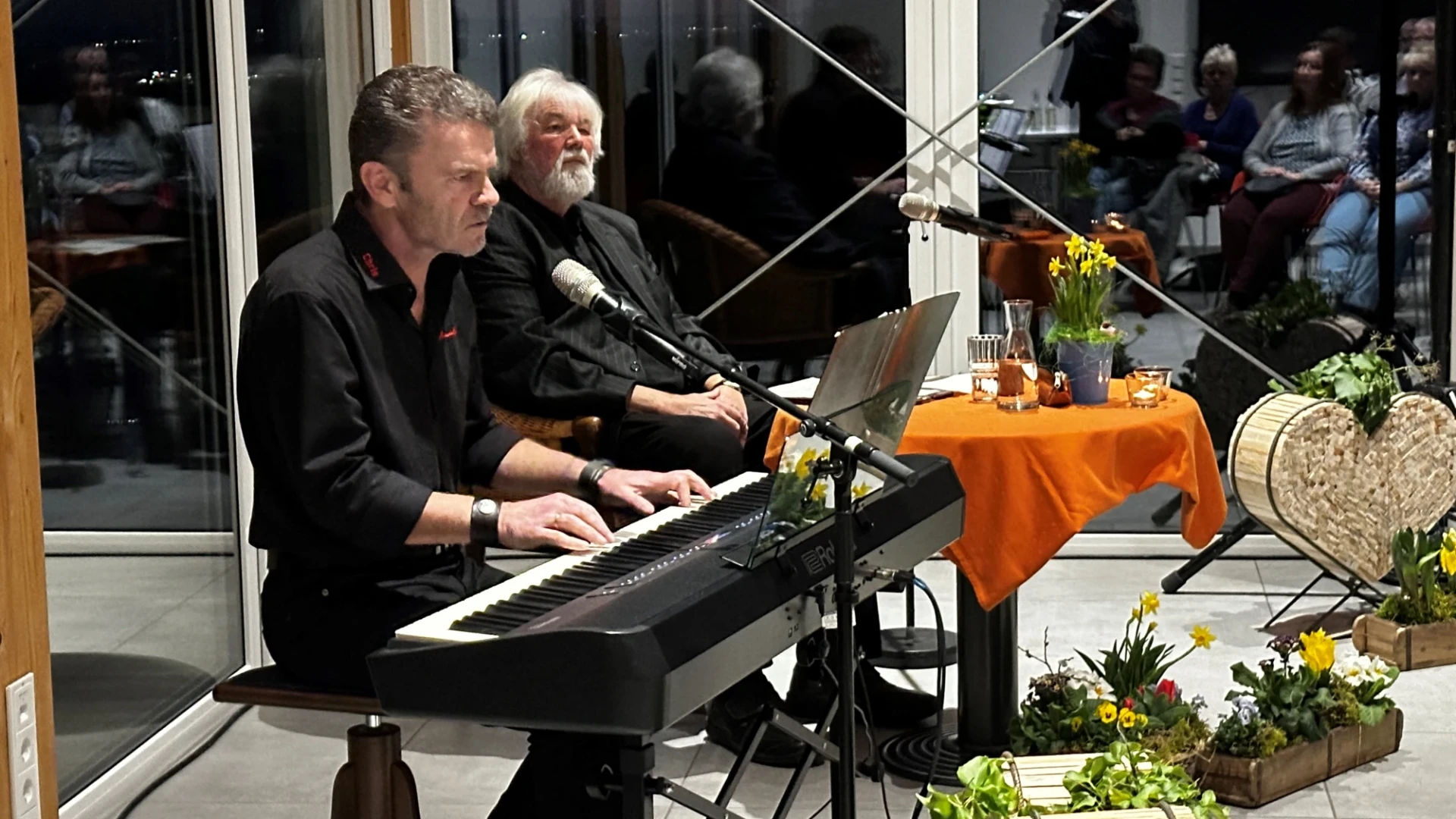 Ein Mann sitzt an einem E-Piano, hinter ihm sitzt ein Mann mit wei&szlig;en Haaren und grauem Bart an einem kleinen runden Tisch. Der Raum hat gro&szlig;e Fenster und ist fr&uuml;hlingshaft dekoriert.