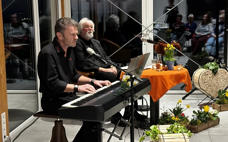 Ein Mann sitzt an einem E-Piano, hinter ihm sitzt ein Mann mit wei&szlig;en Haaren und grauem Bart an einem kleinen runden Tisch. Der Raum hat gro&szlig;e Fenster und ist fr&uuml;hlingshaft dekoriert.