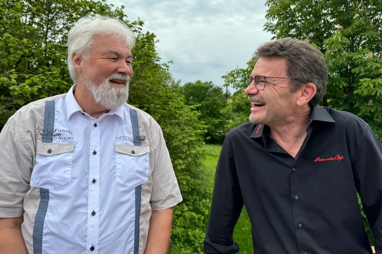 Zwei M&auml;nner stehen sich nebeneinander und l&auml;cheln sich an. Der wei&szlig;haarige Mann tr&auml;gt Bart und hat ein beige-blaues Hemd, der dunkelhaarige Mann tr&auml;gt einen kurzen Bart, eine Brille und ein schwarzes Hemd mit rotem Stickmuster.