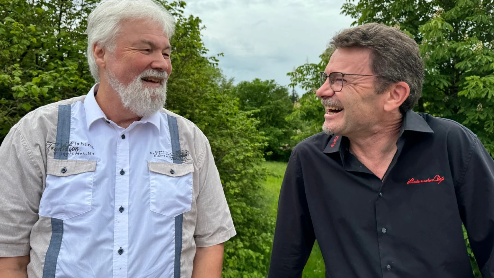 Zwei M&auml;nner stehen sich nebeneinander und l&auml;cheln sich an. Der wei&szlig;haarige Mann tr&auml;gt Bart und hat ein beige-blaues Hemd an. der dunkelhaarige Mann tr&auml;gt einen kurzen Bart, eine Brille und ein schwarzes Hemd mit rotem Stickmuster.