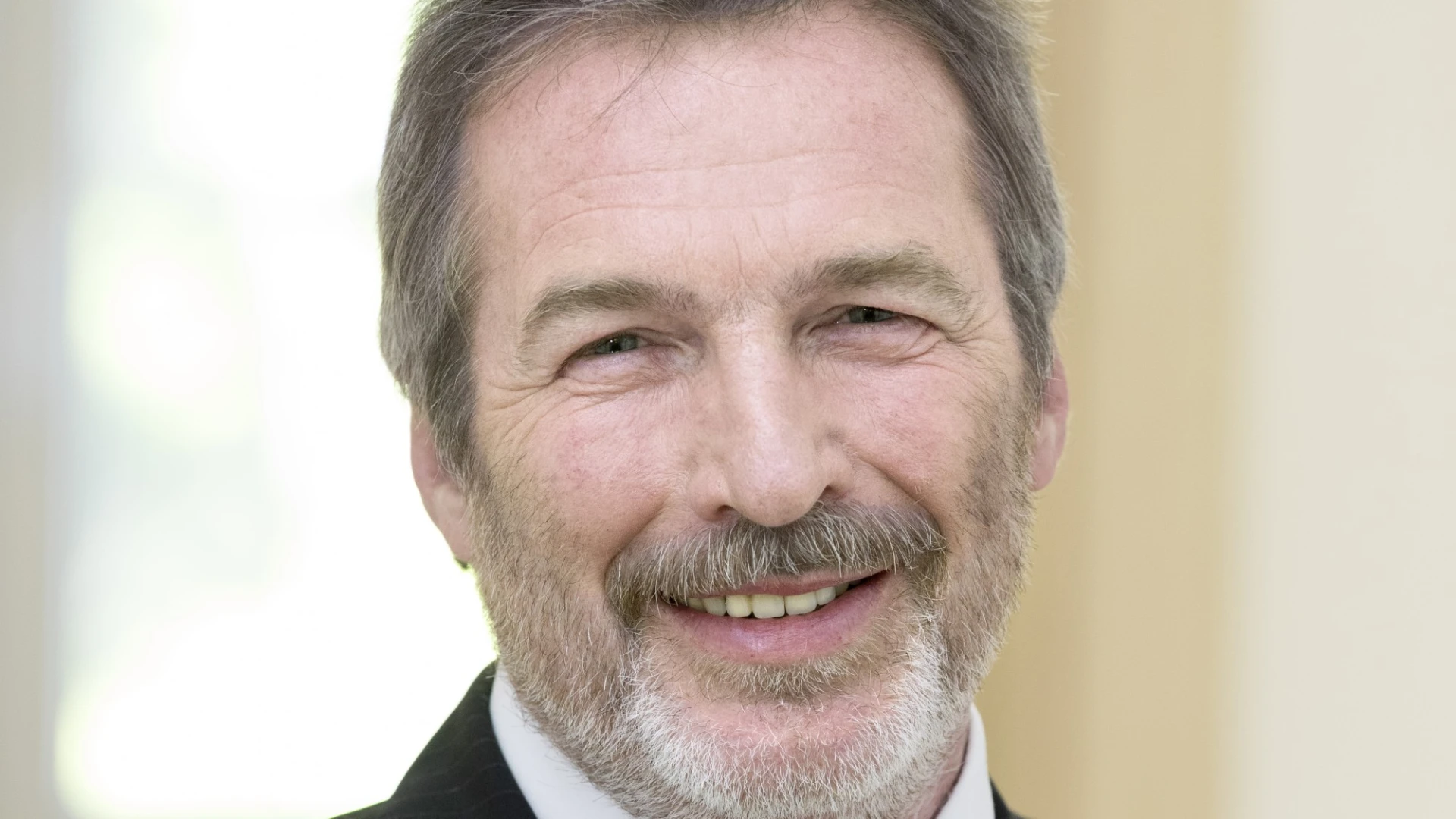 Ein &auml;lterer Mann in einem schwarzen Anzug, Krawatte und wei&szlig;em Hemd ist zu sehen. Er hat graue Haare und einen grauen Bart.