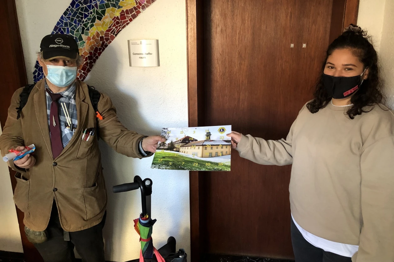 Zwei Frauen halten gemeinsam ein Bild in den H&auml;nden, auf dem zwei gro&szlig;e Geb&auml;ude und eine Wiese zu sehen sind. Sie stehen vor einer Holzt&uuml;r und beide tragen Mund-Nasenschutz.