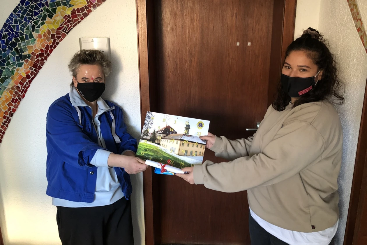 Zwei Frauen halten gemeinsam ein Bild in den H&auml;nden, auf dem zwei gro&szlig;e Geb&auml;ude und eine Wiese zu sehen sind. Sie stehen vor einer Holzt&uuml;r und beide tragen Mund-Nasenschutz.