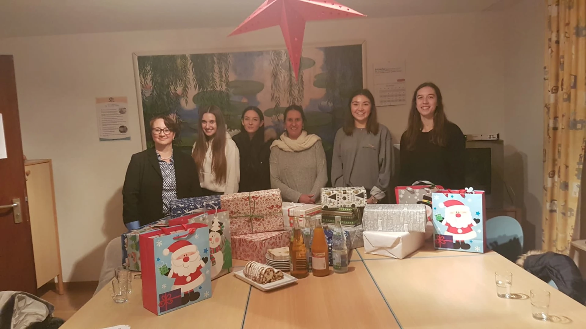 Sechs junge Frauen stehen in einem Raum hinter einem Tisch, auf dem Weihnachtsgeschenke stehen. An der Wand hinter ihnen h&auml;ngt ein gro&szlig;es Bild. Im Vordergrund h&auml;ngt ein gro&szlig;er roter Stern.