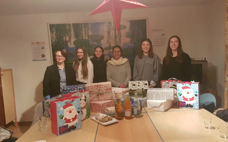 Sechs junge Frauen stehen in einem Raum hinter einem Tisch, auf dem Weihnachtsgeschenke stehen. An der Wand hinter ihnen h&auml;ngt ein gro&szlig;es Bild. Im Vordergrund h&auml;ngt ein gro&szlig;er roter Stern.