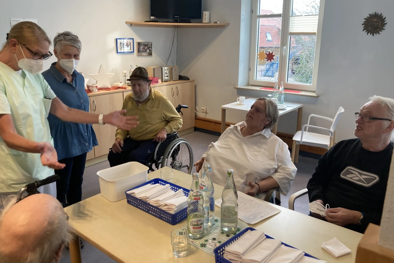 Zwei Frauen mit Mund-Nasen-Schutz stehen bei einer Gruppe &auml;lterer Menschen, die um einen Tisch herumsitzen und zuh&ouml;ren.