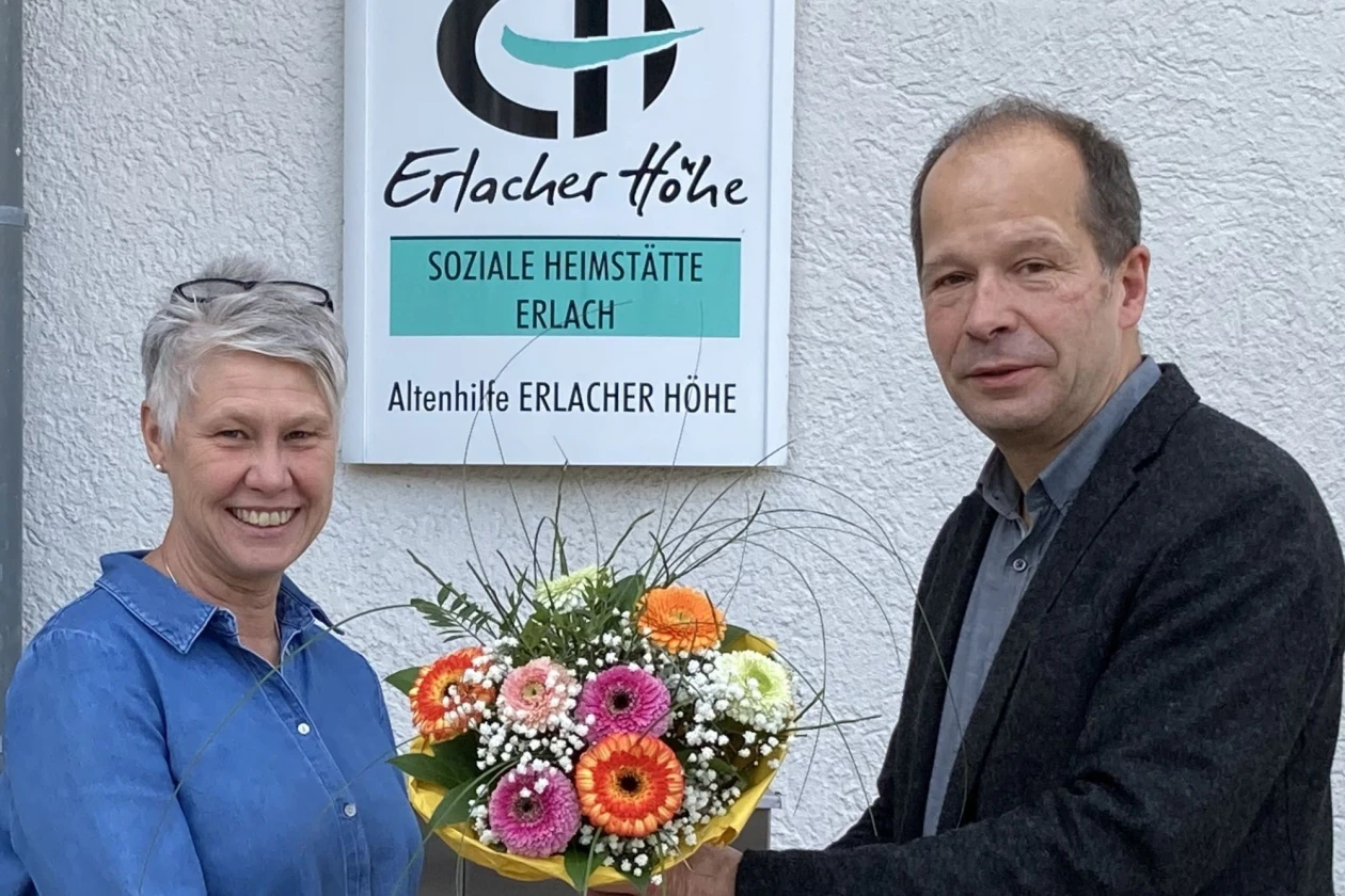 Ein Mann in dunklem Hemd und Jackett &uuml;berreicht einer grauhaarigen Frau mit Brille in den Haaren einen Blumenstrau&szlig;. Beide l&auml;cheln in die Kamera. Im Hintergrund ist ein Schild mit der Aufschrift "Erlacher H&ouml;he Soziale Heimst&auml;tte Erlach" zu sehen.
