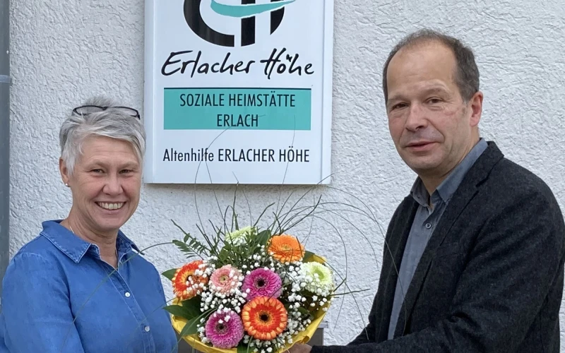 Ein Mann in dunklem Hemd und Jackett &uuml;berreicht einer grauhaarigen Frau mit Brille in den Haaren einen Blumenstrau&szlig;. Beide l&auml;cheln in die Kamera. Im Hintergrund ist ein Schild mit der Aufschrift "Erlacher H&ouml;he Soziale Heimst&auml;tte Erlach" zu sehen.
