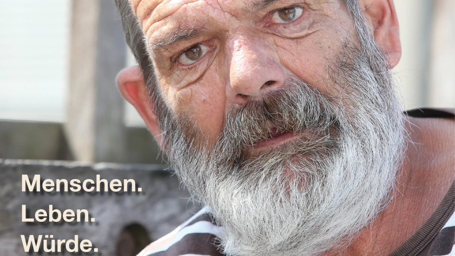 Ein Mann mit grauen Haaren und Bart, blickt in die Kamera, er tr&auml;gt ein gestreiftes T-Shirt. Im Hintergrund ist ein Gel&auml;nder und unten links eine Texteinblendung: "Menschen. Leben. W&uuml;rde.