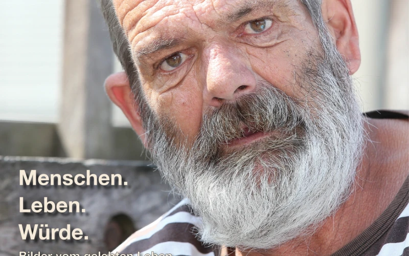 Ein Mann mit grauen Haaren und Bart, blickt in die Kamera, er tr&auml;gt ein gestreiftes T-Shirt. Im Hintergrund ist ein Gel&auml;nder und unten links eine Texteinblendung: "Menschen. Leben. W&uuml;rde.