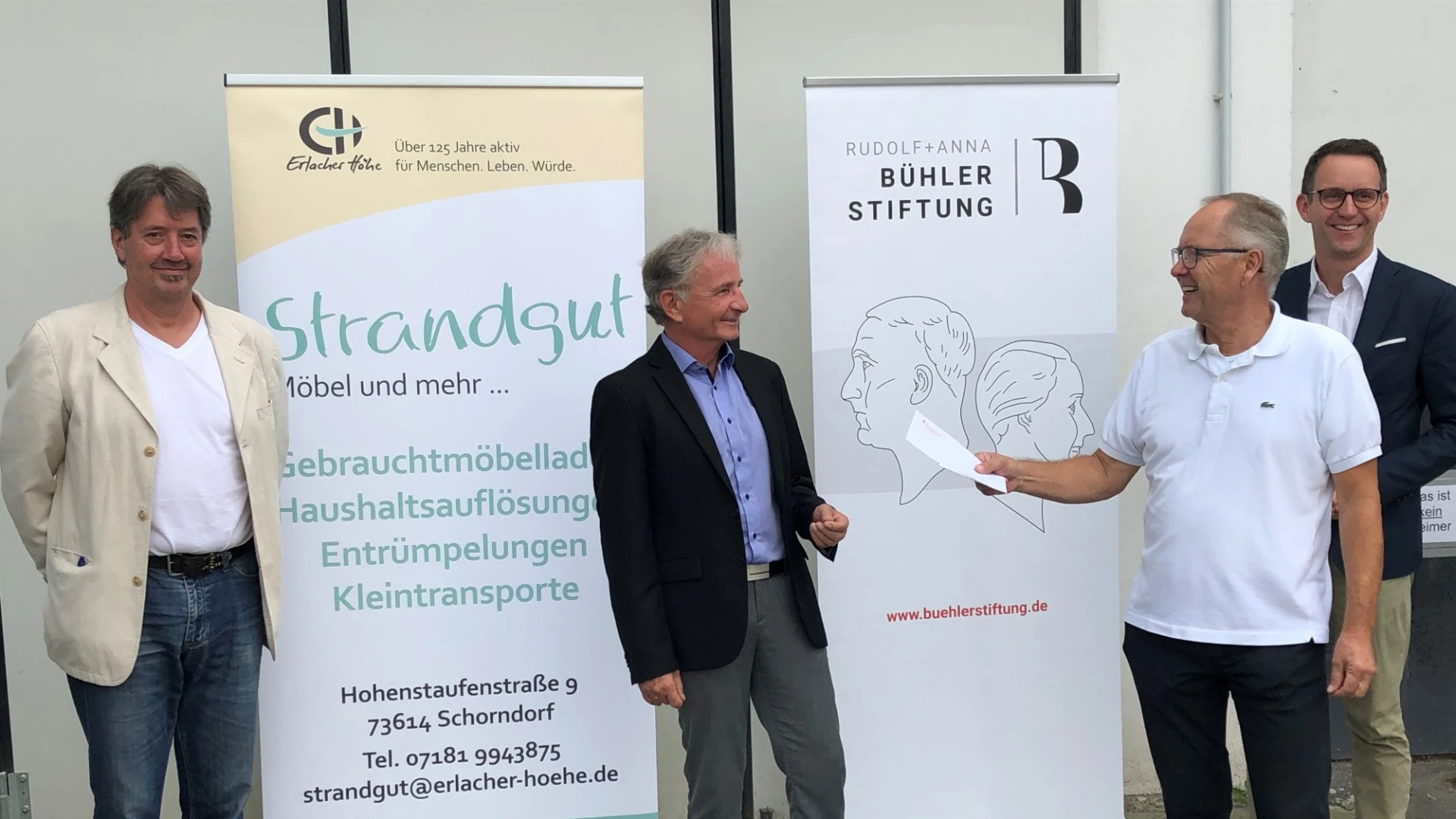 Vier M&auml;nner stehen vor zwei Infobannern mit den Aufschriften "Strandgut" und "B&uuml;hler Stiftung". Der Mann im Vordergrund auf der rechten Seite h&auml;lt ein Papier in der Hand.